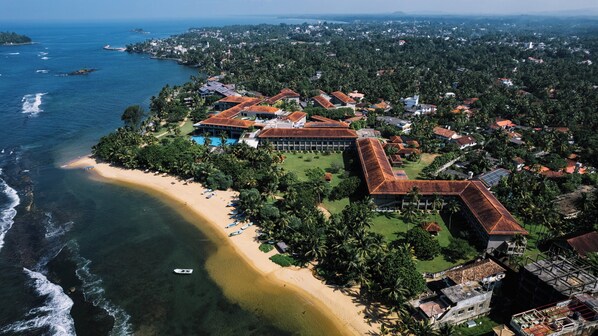 Vista aérea de un complejo turístico costero con playa, palmeras y un edificio grande de techo rojo.