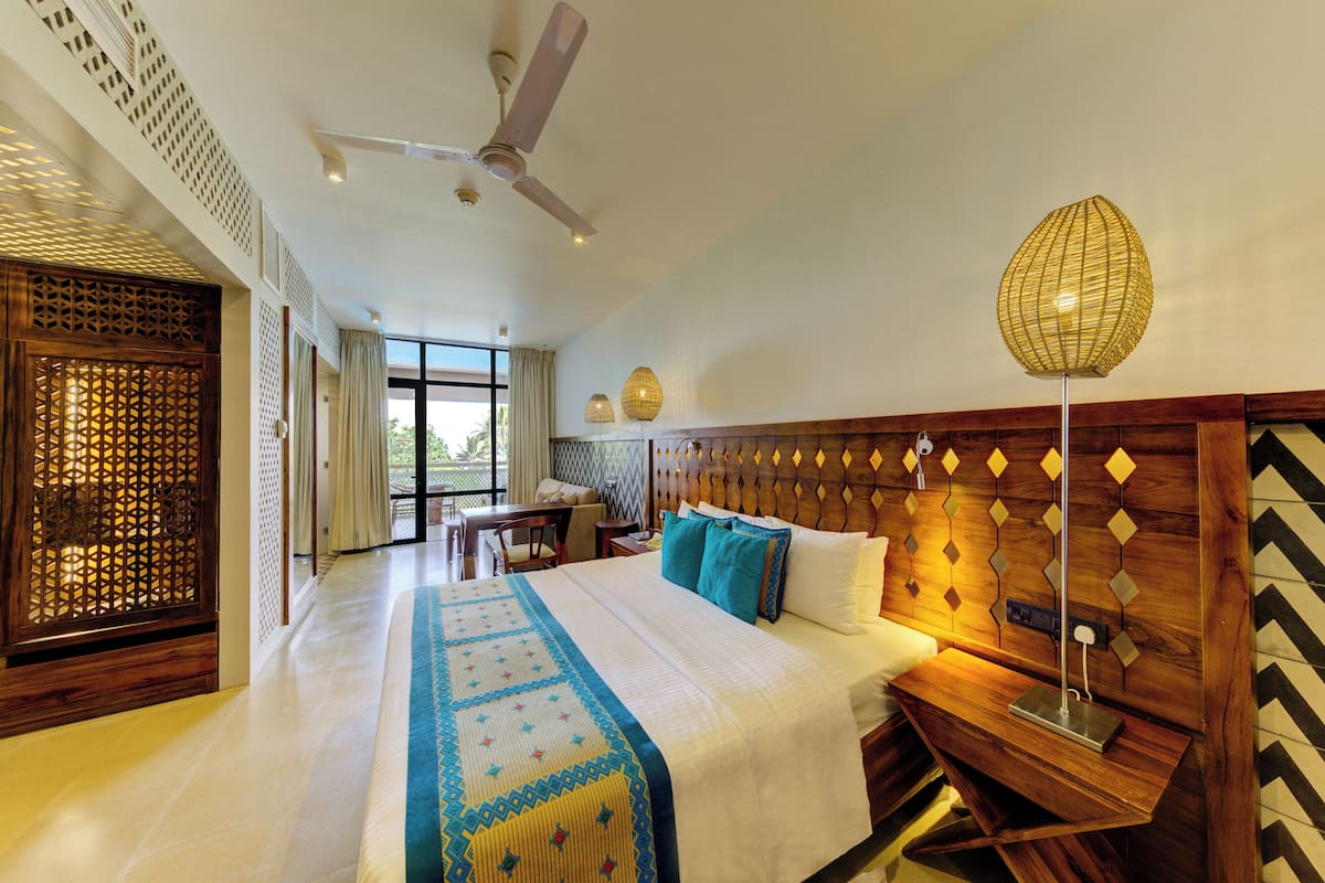 Quarto de hotel com cama, escrivaninha, cadeira, ventilador de teto e uma janela grande.