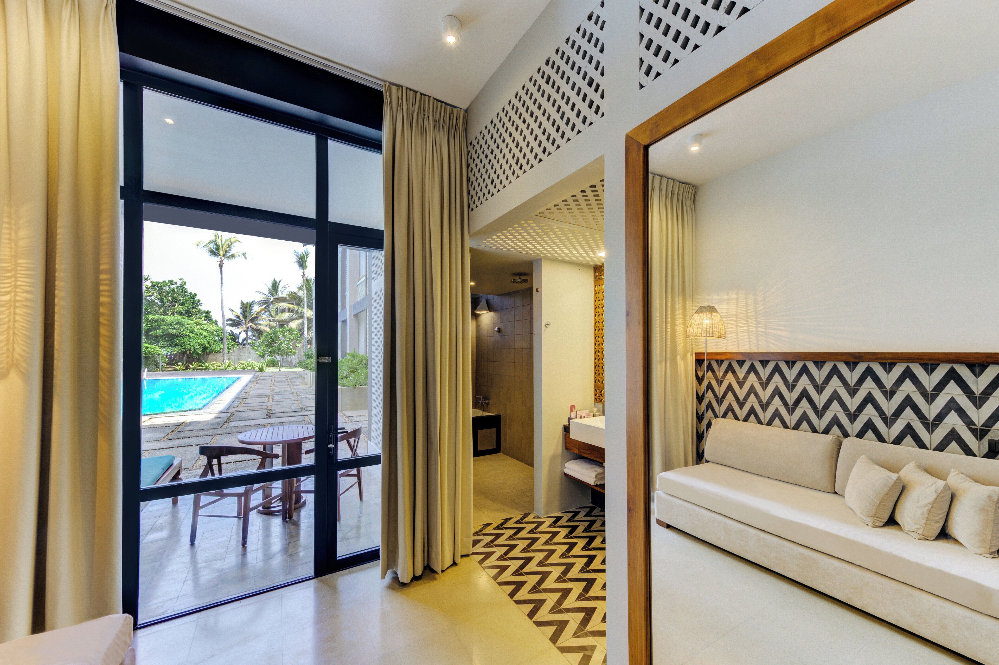 A modern hotel room with a large sliding glass door leading to a pool area, a sofa, and a bathroom with a vanity.