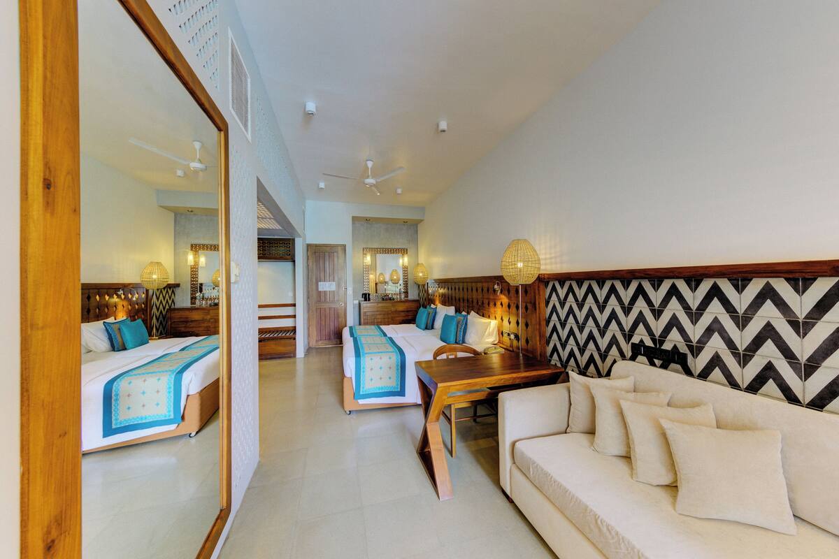 Quarto de hotel com duas camas, um sofá, uma mesa de madeira e parede com padrão Chevron.