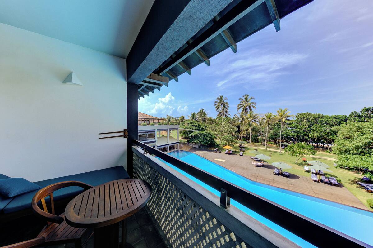 Varanda com mesa e cadeiras de madeira, vista para uma piscina e área ajardinada com palmeiras.