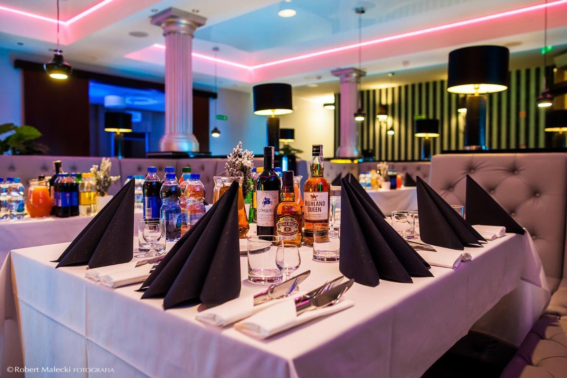 A restaurant table set with multiple bottles of alcohol, glasses, and cutlery, with a modern interior and ambient lighting.