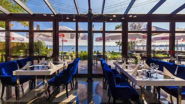 An outdoor dining area with tables and blue chairs, large umbrellas, and a view of the water.