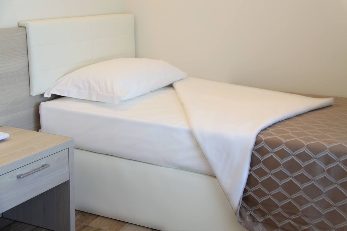 A neatly made bed with a white and brown patterned bedspread, a white pillow, and a wooden nightstand with a drawer.