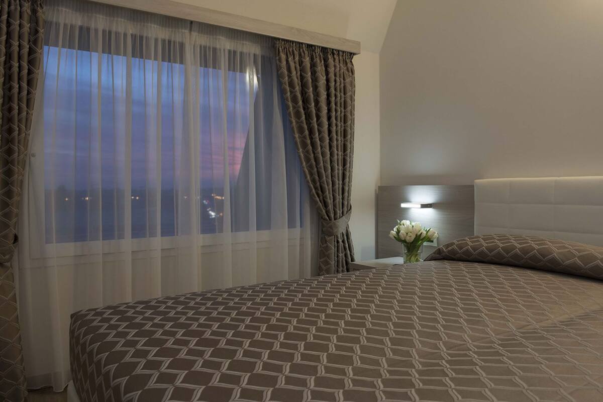 A hotel room with a large bed, a nightstand with a lamp, and a view of the city at dusk.