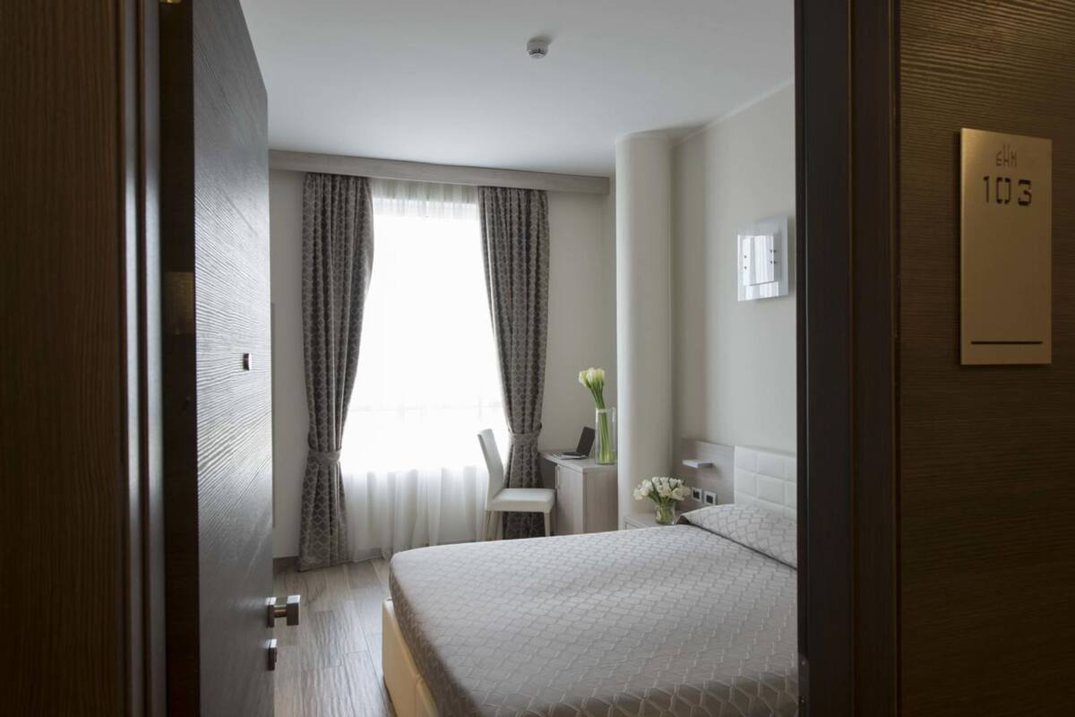 A hotel room with a bed, a desk, and a chair. There is a window with curtains, a vase with flowers, and a sign with the room number.