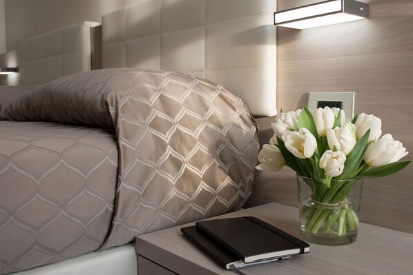 Una cama bien hecha con una almohada estampada, una mesita de noche con un libro y un jarrón de flores blancas, y un aplique de pared.