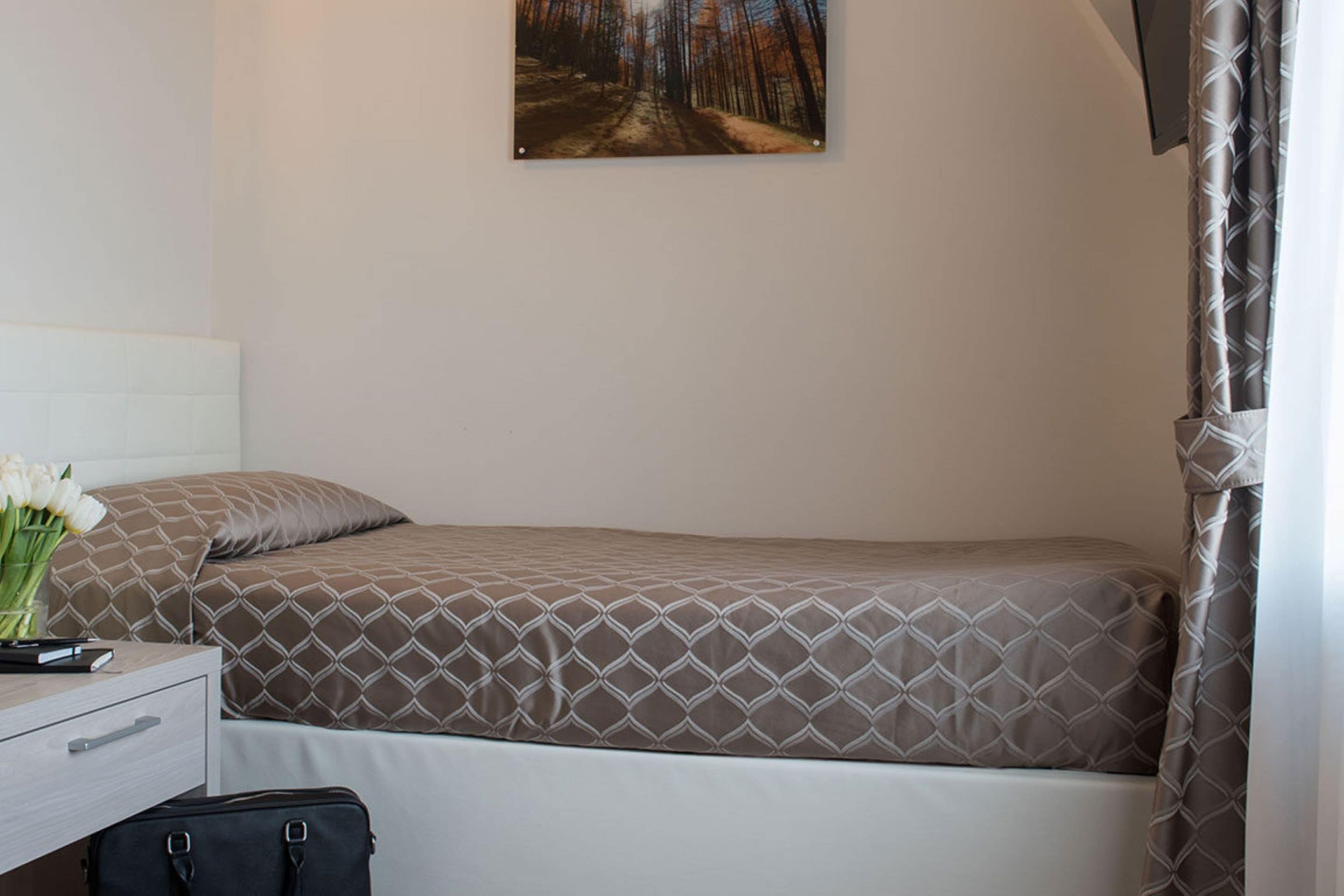 A single bed with a patterned bedspread, a bedside table with a lamp, a small desk with a chair, and a painting on the wall.