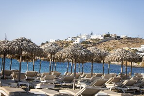 A beach with rows of sun loungers and thatched umbrellas, a clear blue sea, and white buildings on a hillside in the background.