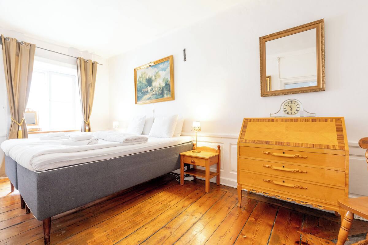 A bedroom with a bed, a wooden dresser, a nightstand with a lamp, a painting on the wall, and a mirror.