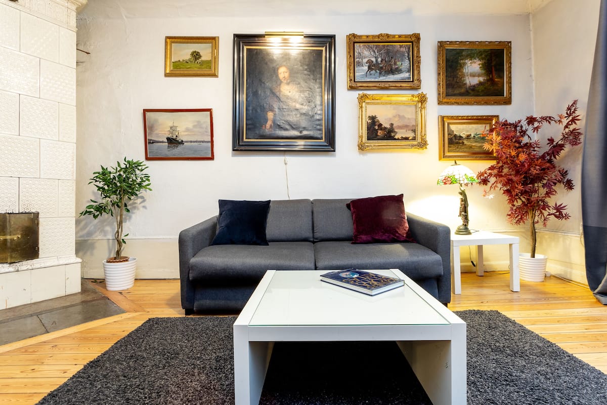 A living room with a grey sofa, a white coffee table, and a fireplace. There are framed pictures on the wall and a potted plant on the floor.