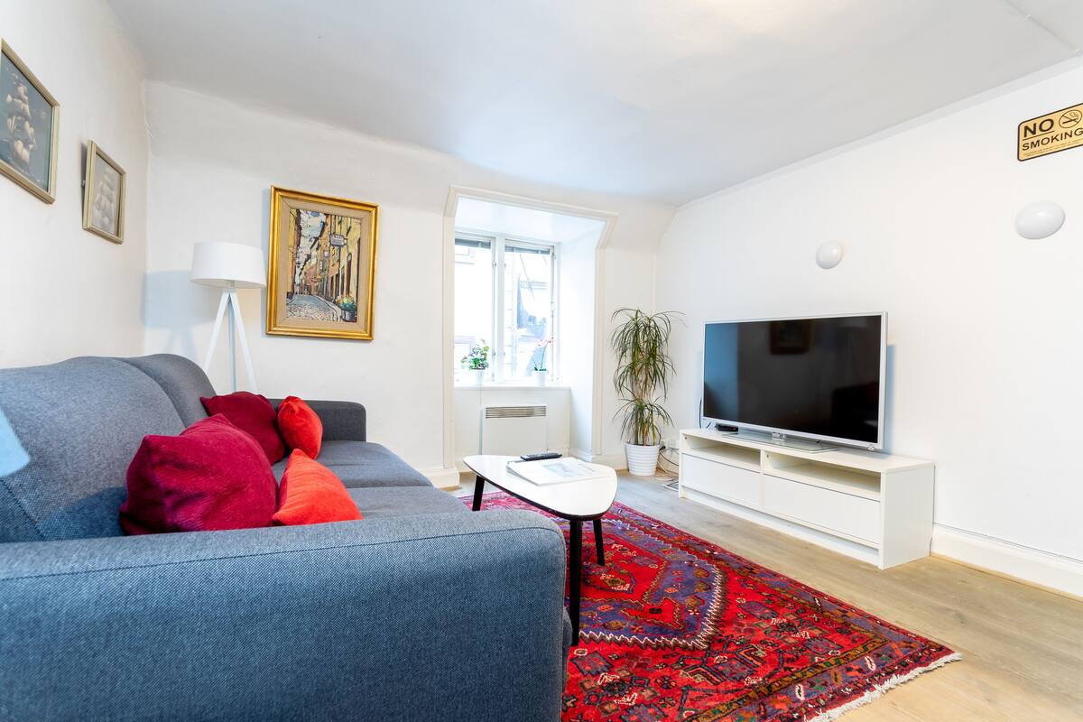 A modern living room with a grey sofa, red cushions, a white coffee table, a large flat-screen TV, and a red patterned rug.