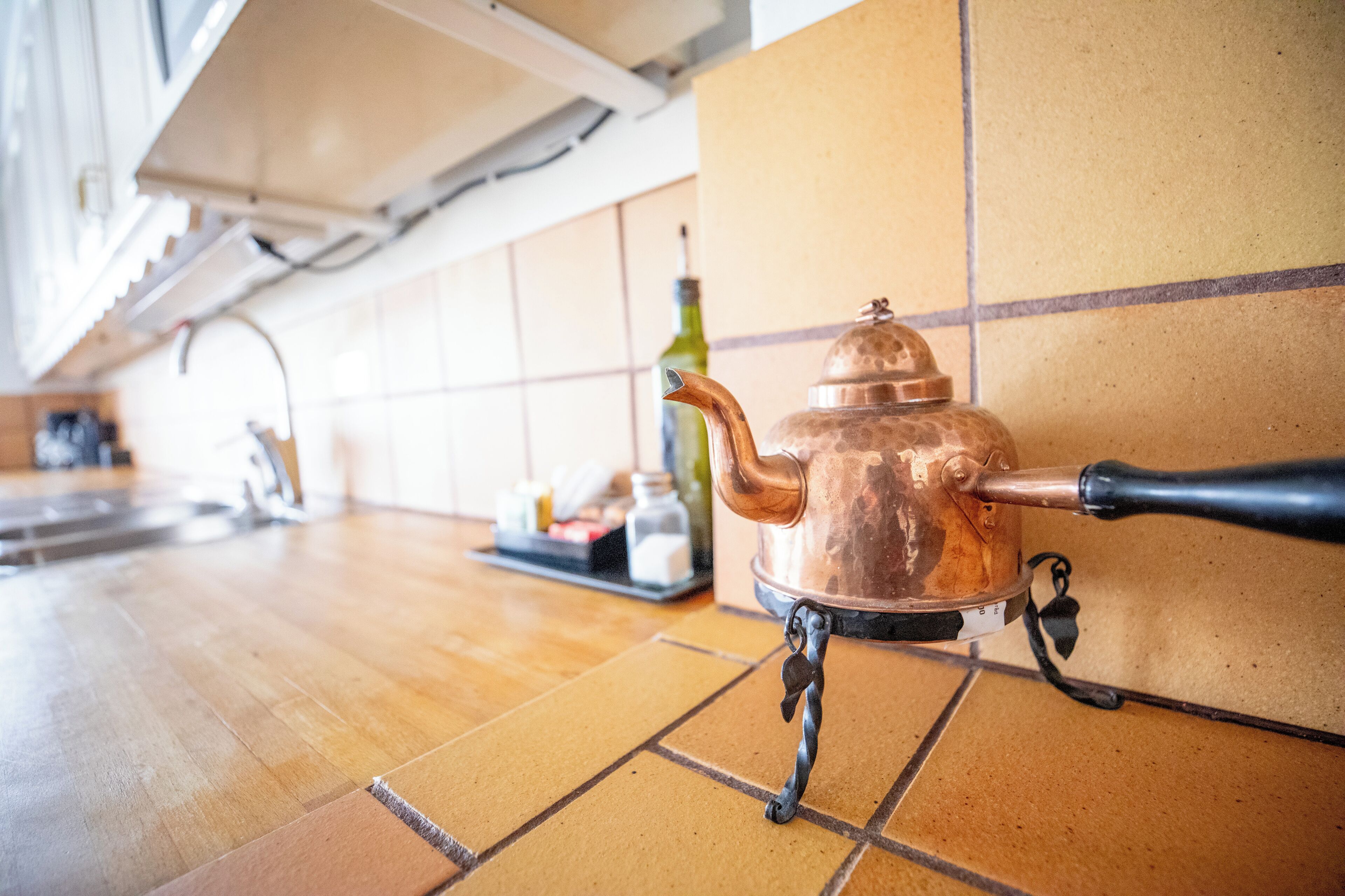 A copper teapot on a tiled kitchen counter with a bottle and other items in the background.