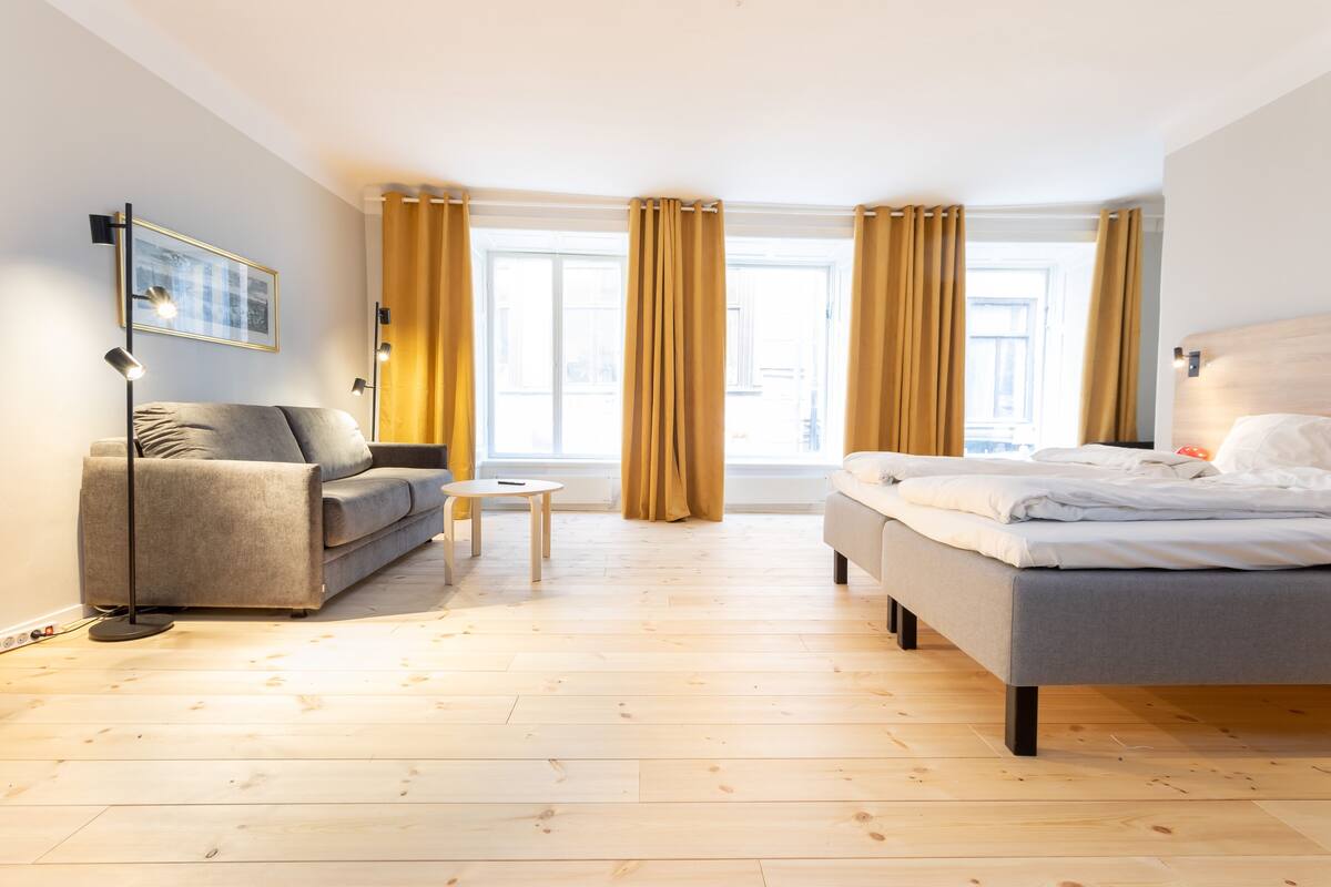 A modern hotel room with a grey sofa, a small round table, a bed with white bedding, and large windows with curtains.