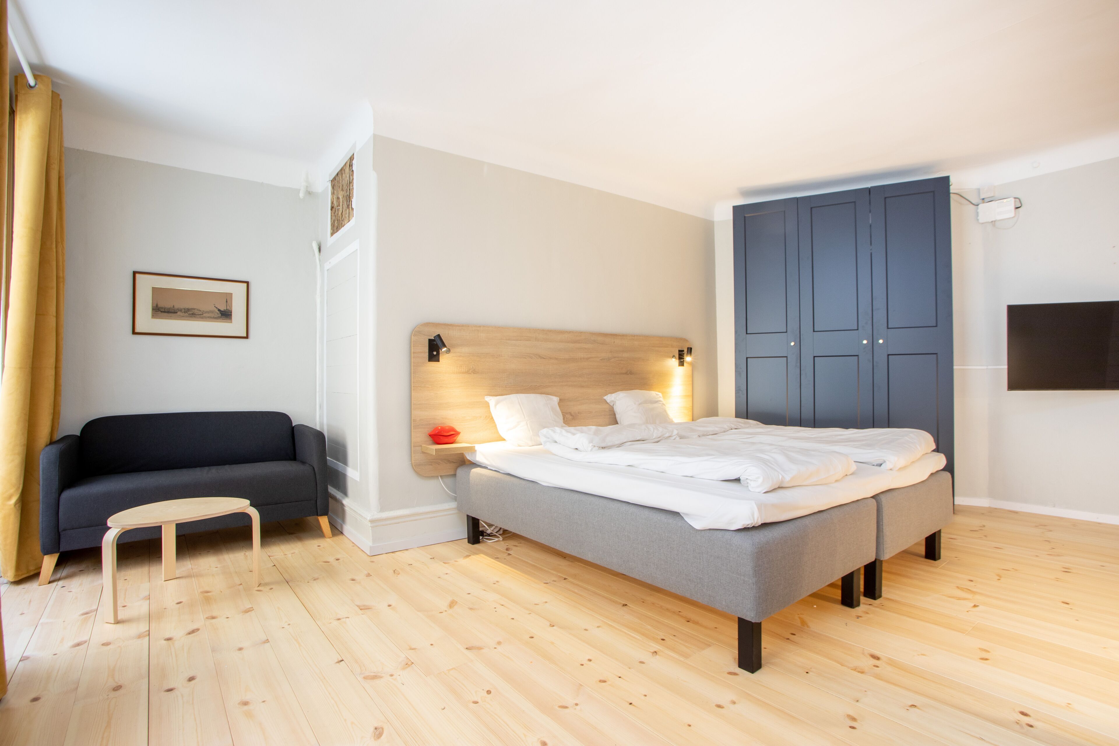 A bedroom with a bed, a sofa, a wooden floor, a wall-mounted TV, and a door.