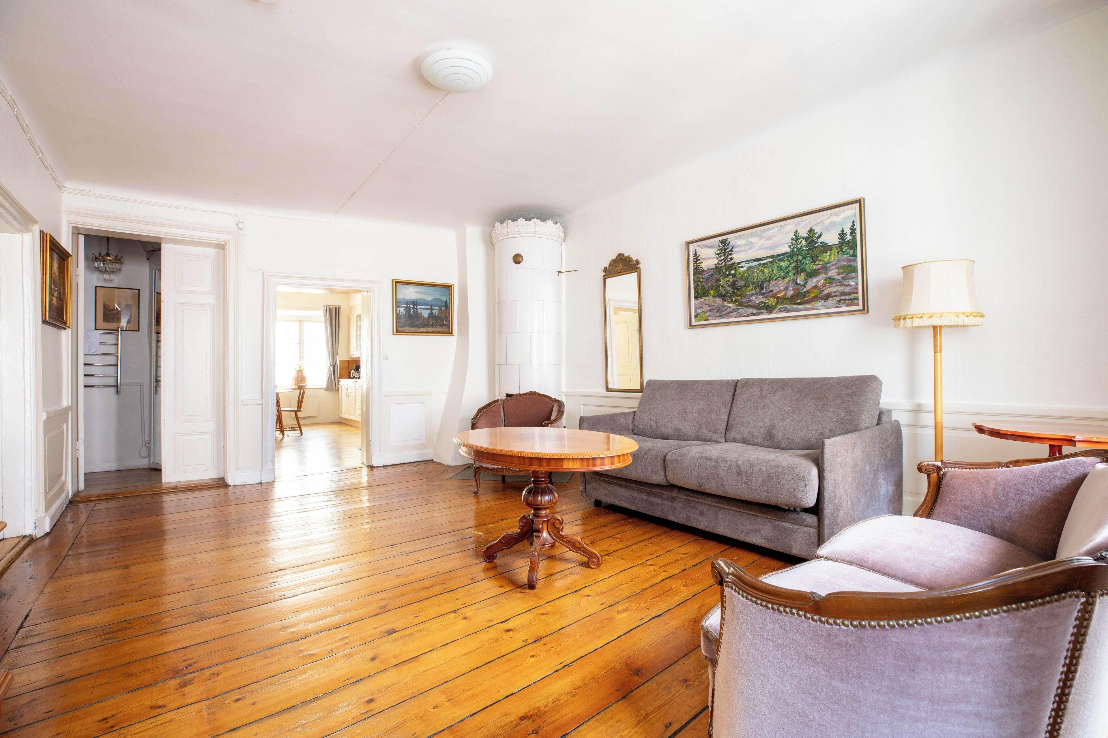 A spacious living room with a wooden floor, a round wooden table, a grey sofa, and a chair. There is a painting on the wall and a lamp on a side table.
