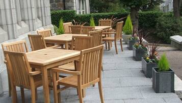 An outdoor patio with wooden tables and chairs, surrounded by potted plants and a stone wall.