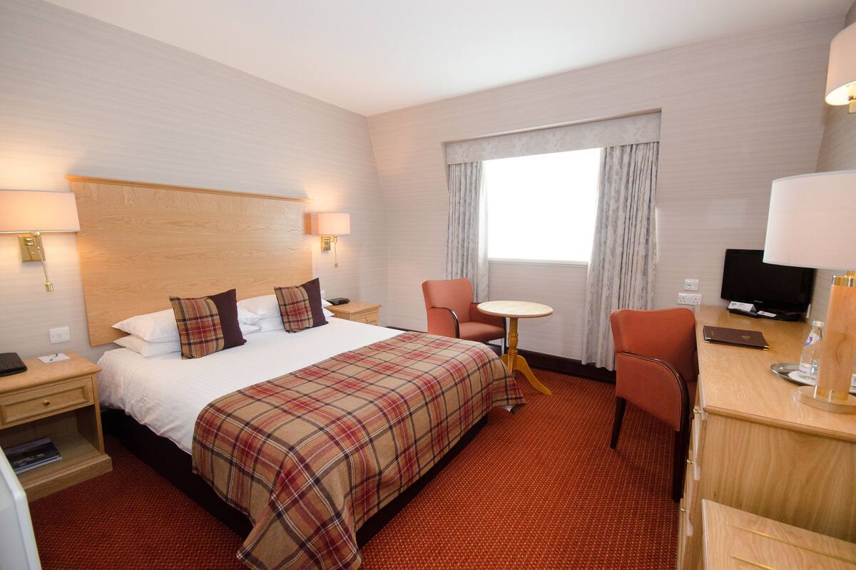 A hotel room with a bed, two chairs, a desk, a television, and a window with curtains.
