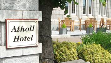 A sign for the Atholl Hotel, a stone building with a tree in front, outdoor seating, and a well-maintained garden.