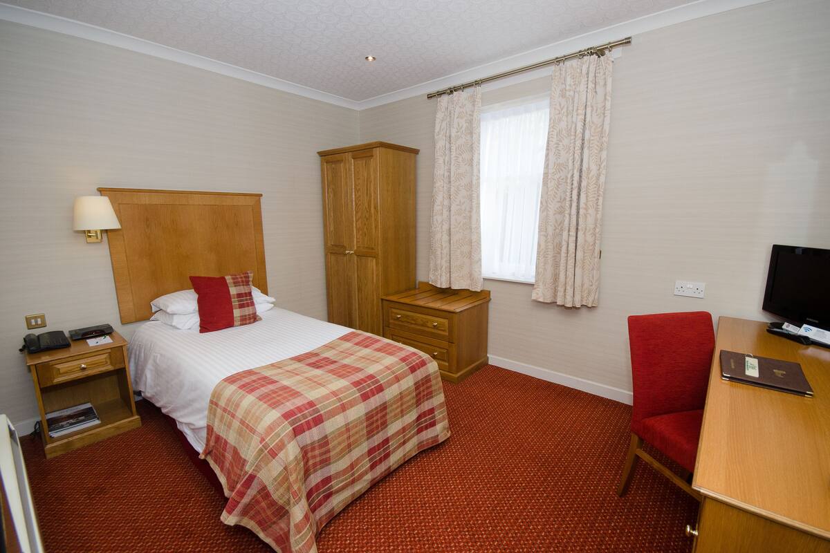 A hotel room with a bed, a wooden wardrobe, a desk with a chair, a television, and a window with curtains.