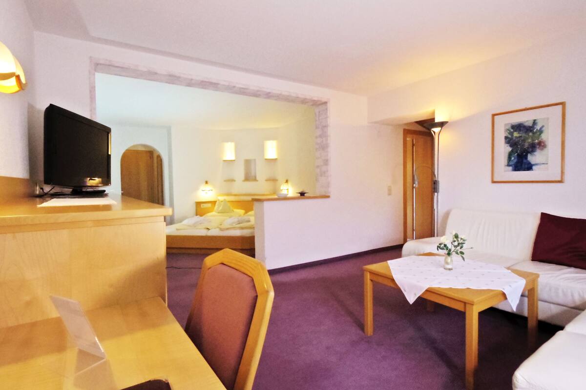 Ein Hotelzimmer mit einem Holztisch, einem Fernseher, einem Bett mit weißem Kopfteil, einer weißen Couch, einem kleinen Tisch mit einer Vase und einem gerahmten Bild an der Wand.