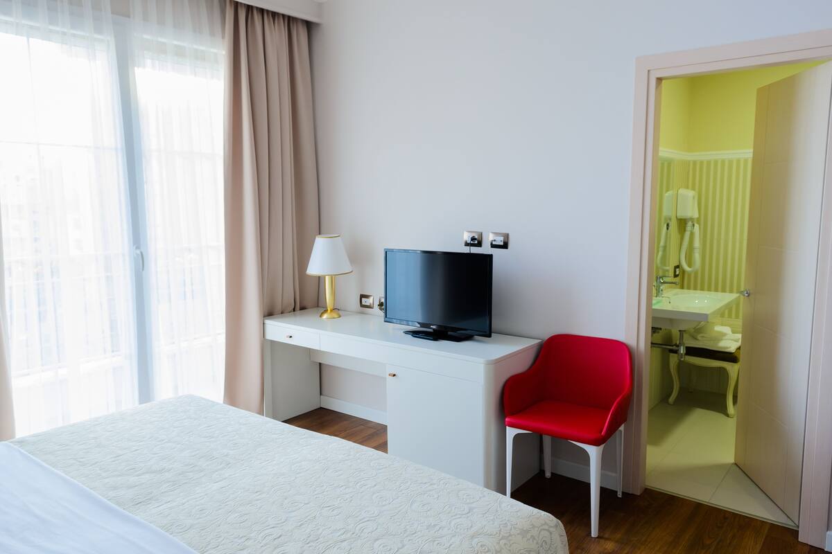 A hotel room with a bed, a desk with a television, a red chair, and a bathroom visible through an open door.