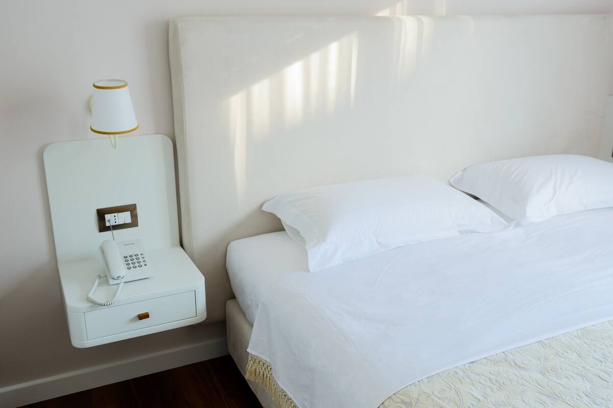 A hotel room with a bed, a nightstand with a phone, and a wall-mounted lamp.