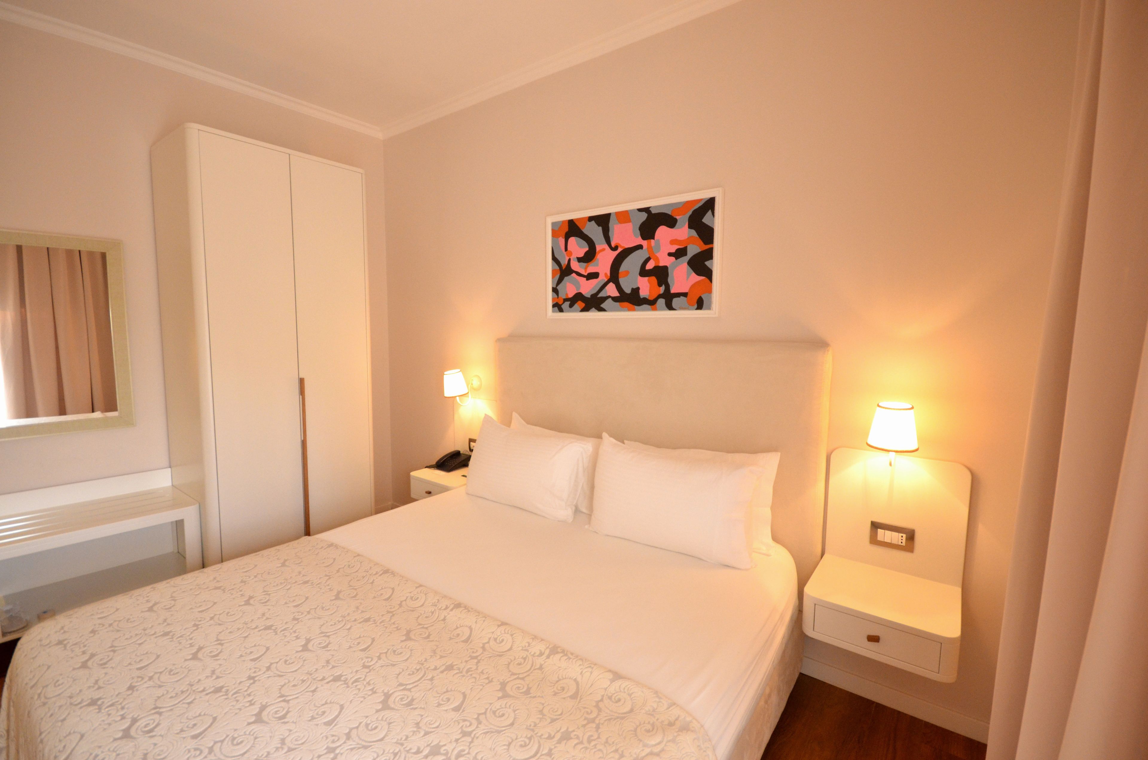 Una cama bien hecha con sábanas blancas y una colcha estampada, dos mesitas de noche con lámparas, una mesita con un teléfono y una puerta a la izquierda.