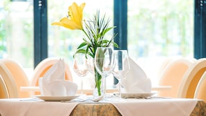 Mesa de comedor con mantel blanco, copas de vidrio transparente y un centro de mesa con una flor amarilla y follaje verde.