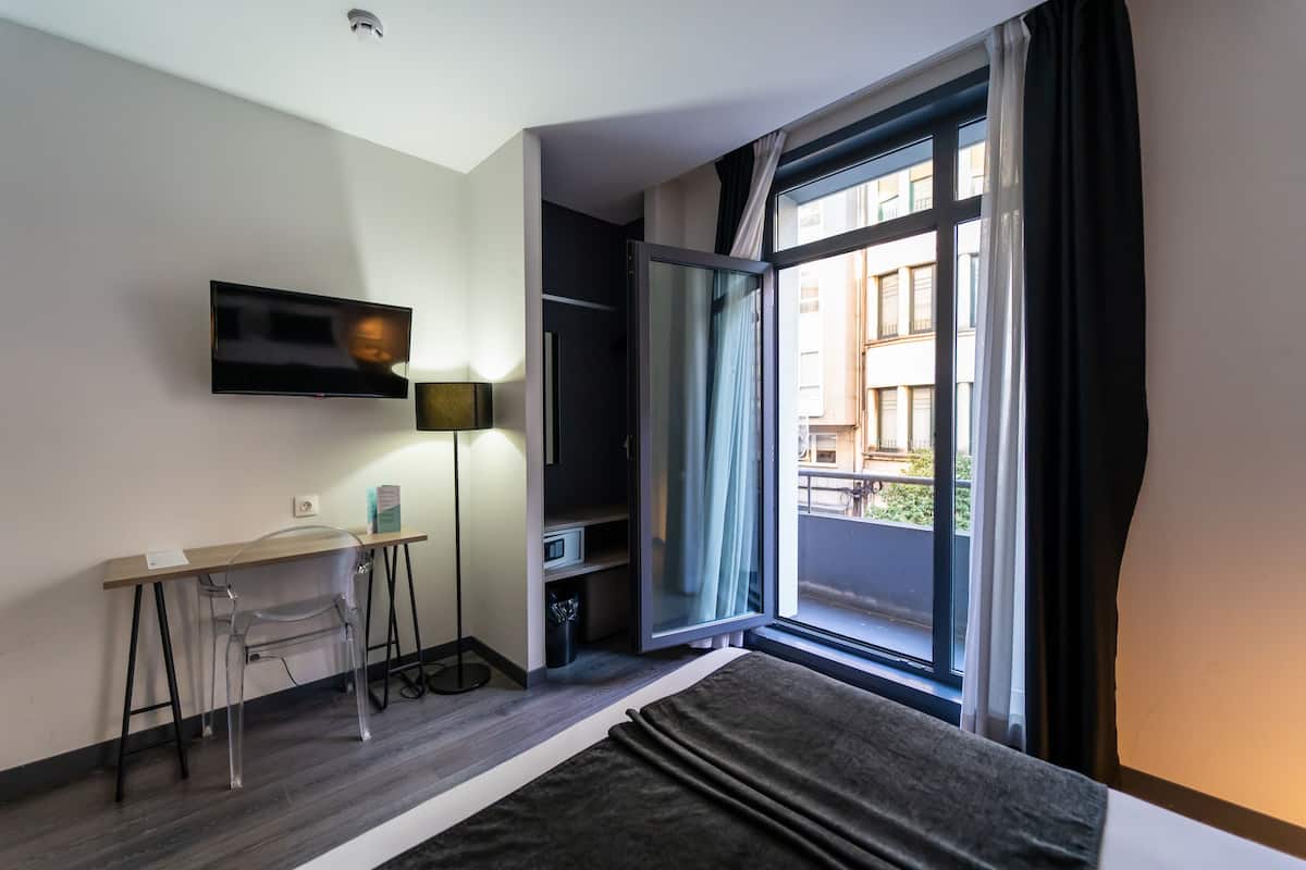 A modern hotel room with a bed, a desk with a chair, a TV mounted on the wall, and a large window with a view of the outside.