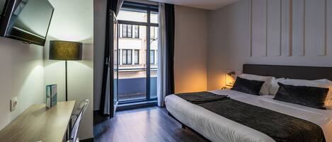 A hotel room with a bed, a desk, a chair, a TV, and a window with a view of buildings.