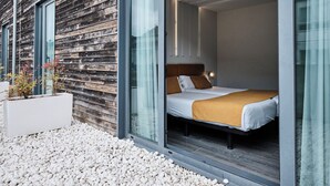 Una habitación de hotel moderna con fachada de madera, grandes puertas correderas de vidrio, cama con ropa de cama blanca y marrón, y un pequeño balcón con guijarros blancos.