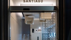 La entrada a un hotel llamado 'LUX SANTIAGO', con un diseño moderno que incluye una puerta de vidrio y una recepción visible.