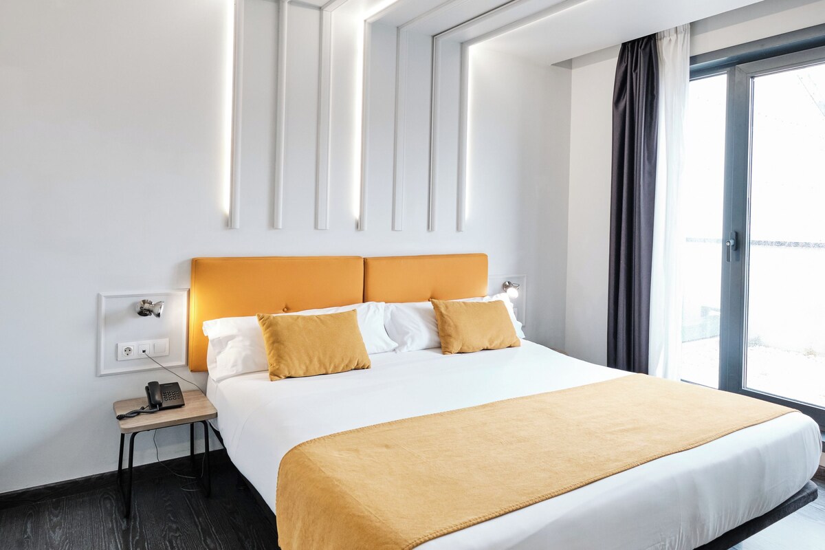 A modern hotel room with a large bed, a bedside table with a phone, and a window with curtains.