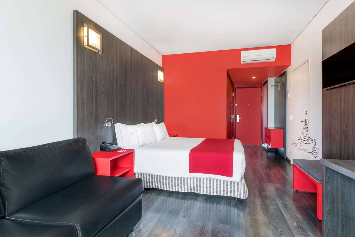 A hotel room with a black leather sofa, a bed with white bedding and a red accent, a red side table, a wooden wall panel, and a TV on the right.