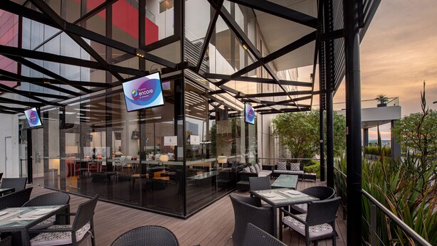 Un patio al aire libre con mobiliario de mimbre y divisiones de vidrio, que muestra señalización con 'encore' y 'encore by'.
