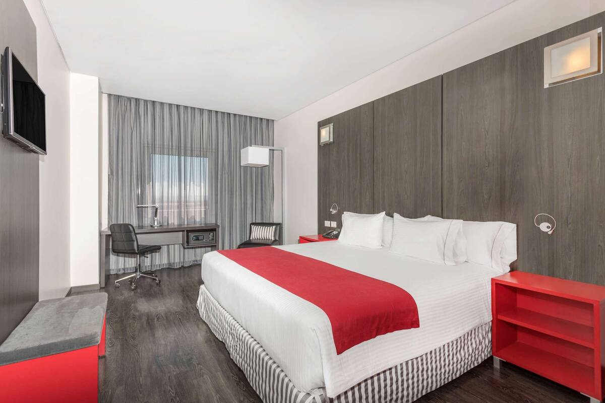 A modern hotel room with a large bed, a desk with a chair, a TV mounted on the wall, and a red nightstand.