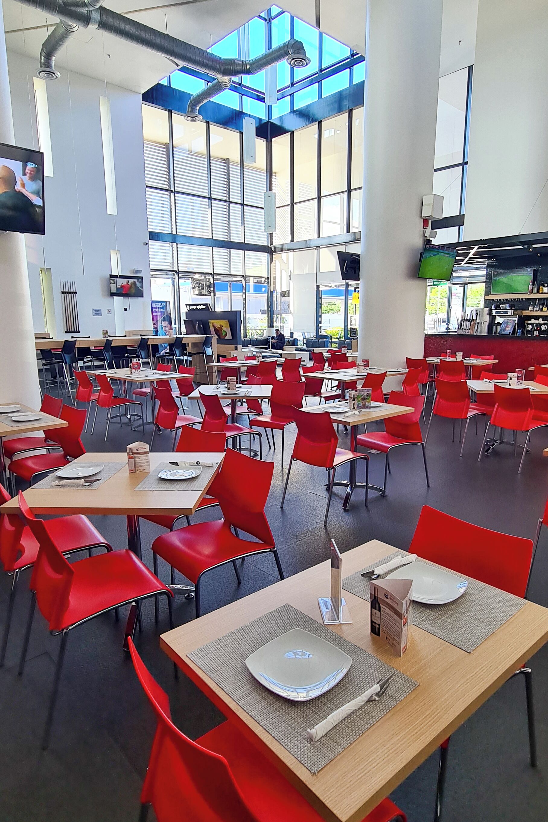 Una amplia cafetería con sillas y mesas rojas, ventanas grandes y una pantalla de televisión.