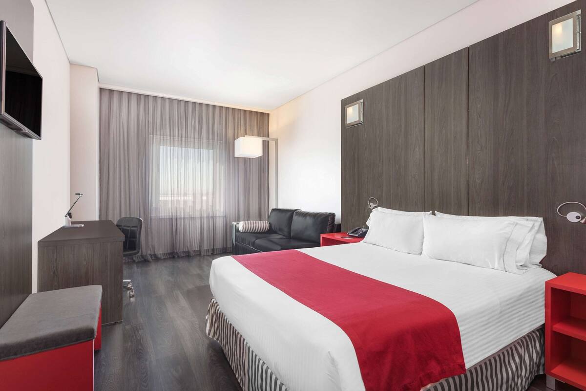 A hotel room with a large bed, a desk, a chair, and a TV mounted on the wall.