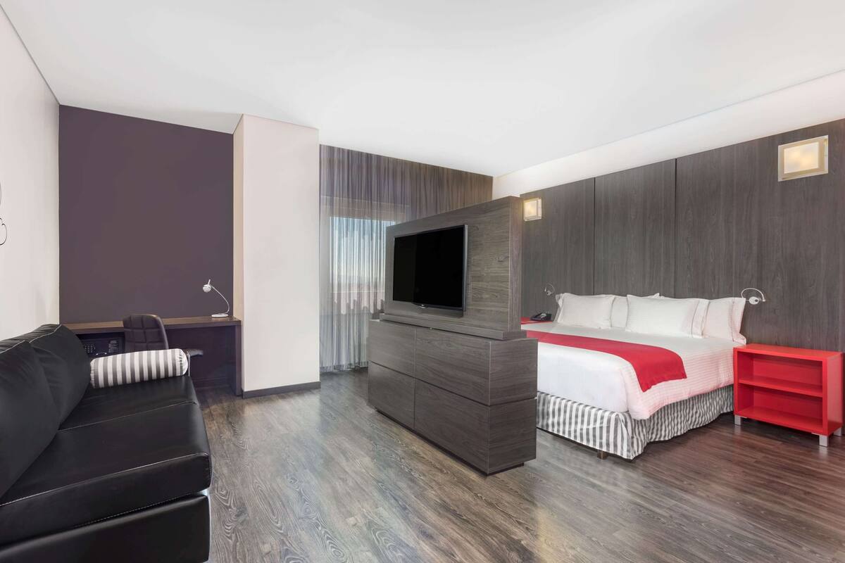 A modern hotel room with a flat-screen TV mounted on a dark wood cabinet, a black leather sofa, a bed with white bedding and a red accent, and a red side table.
