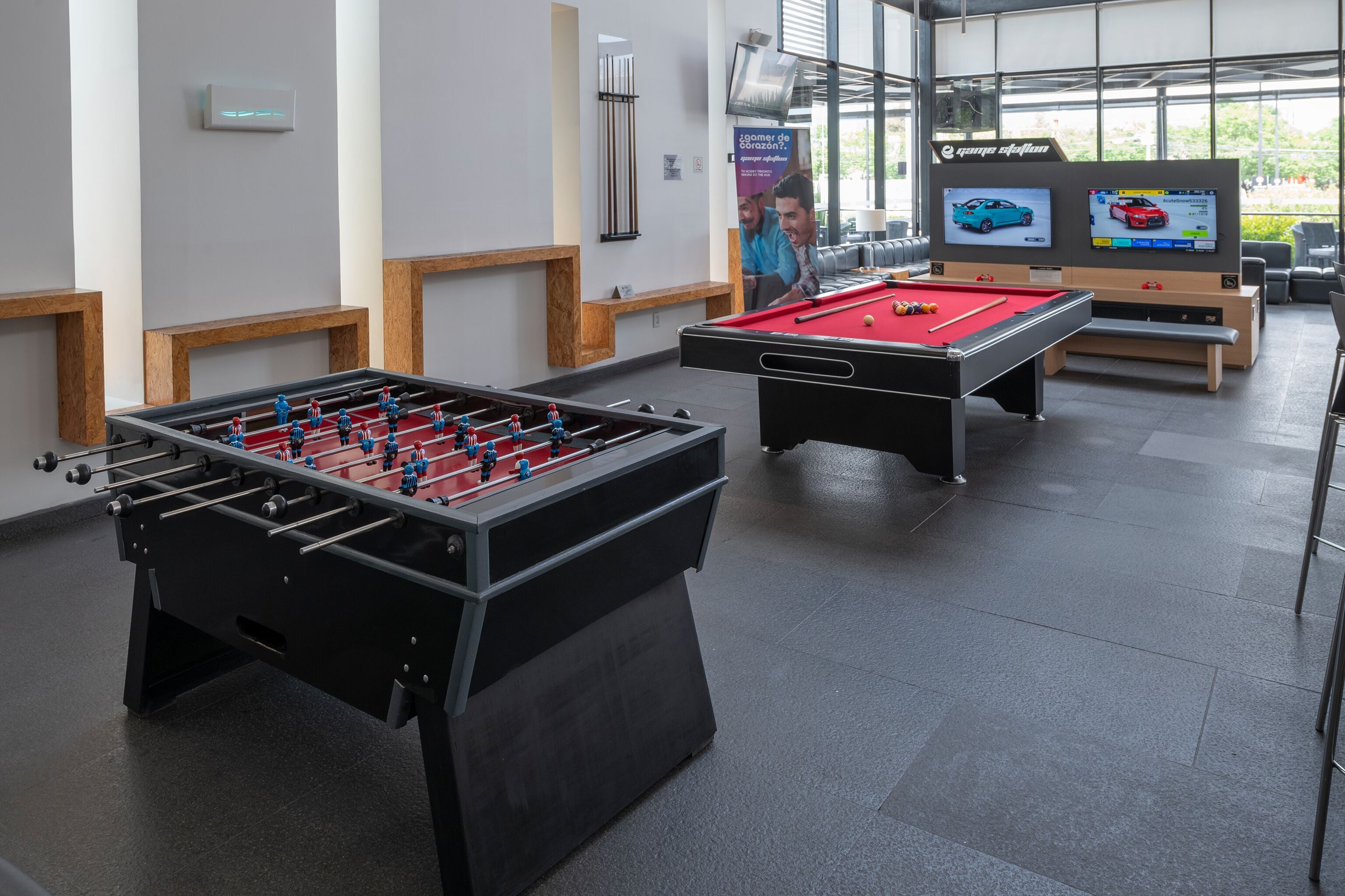 A modern indoor recreational area with two foosball tables and a pool table, featuring a clean, minimalist design with wooden accents and large windows.