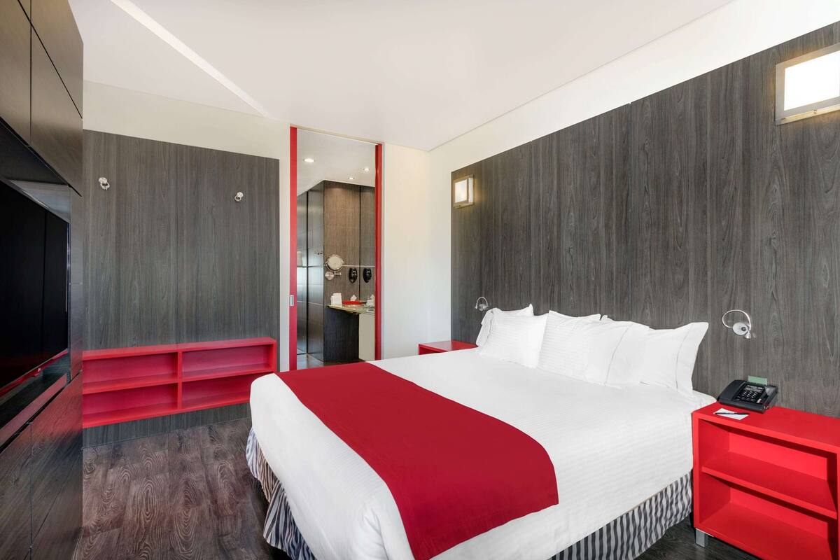 A modern hotel room with a large bed, a red nightstand, a red bench, and a mirror.