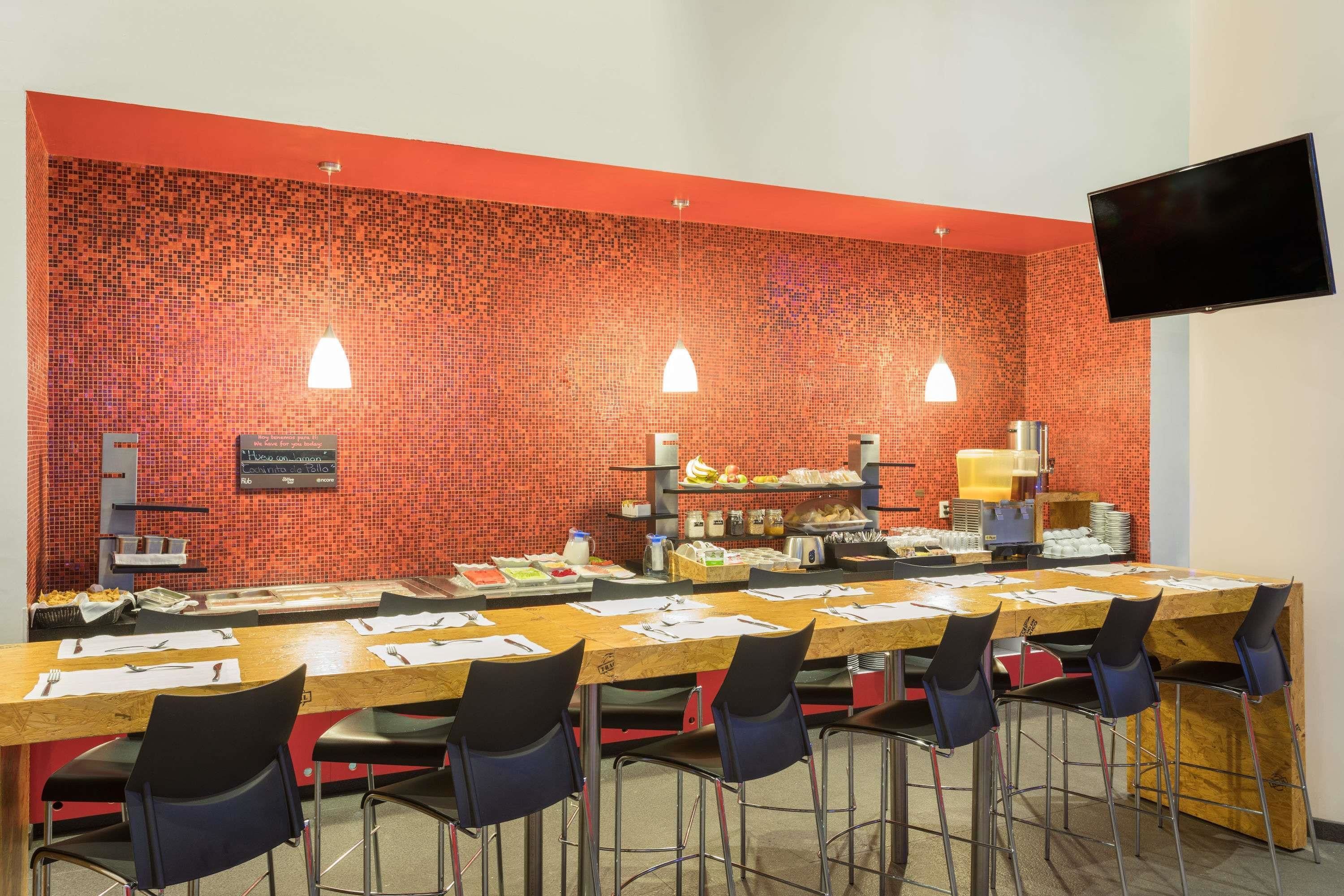 A conference room with a long table, multiple chairs, a flat-screen TV mounted on the wall, and a red-tiled wall.