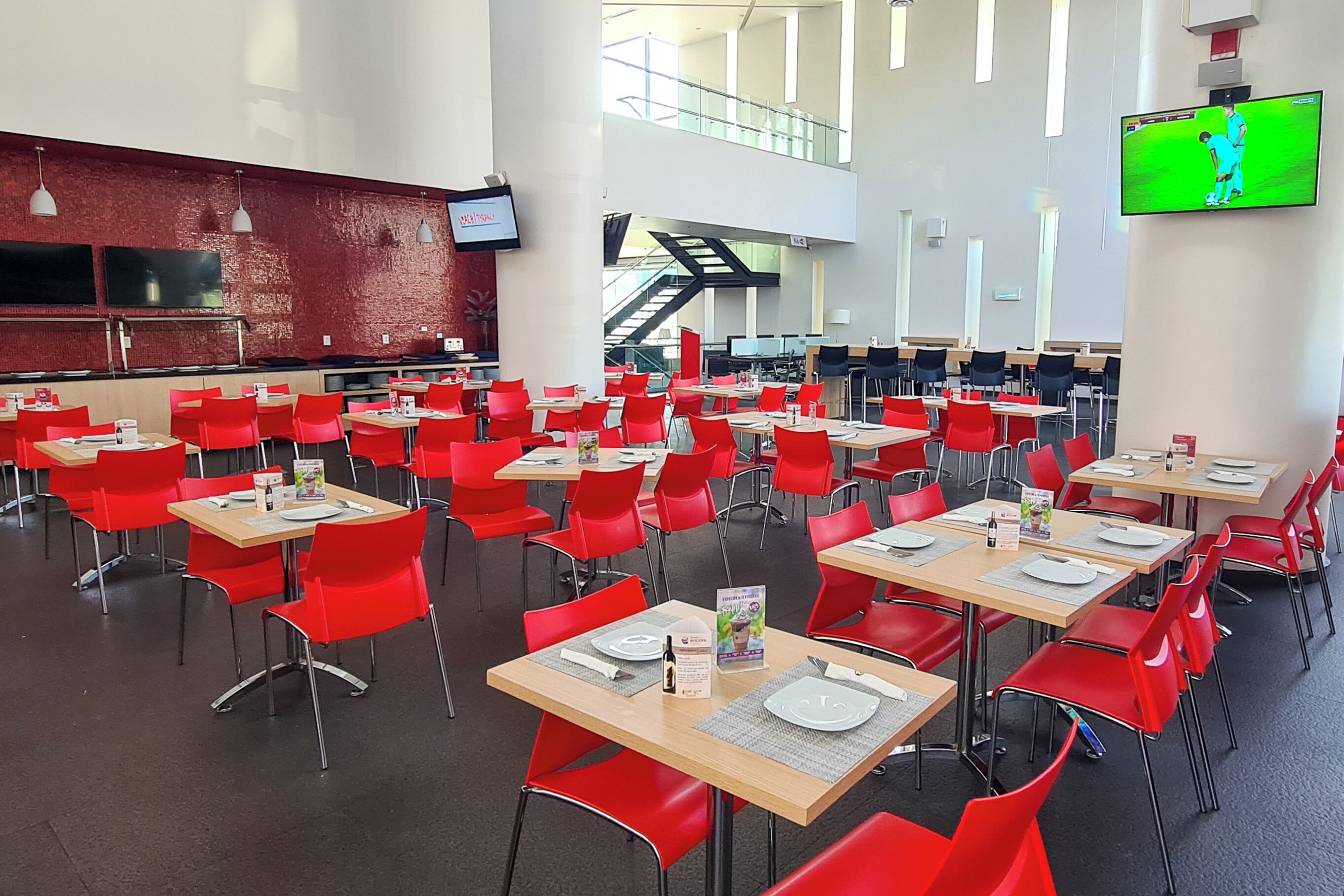 A spacious cafeteria with red chairs and tables, a television screen, and a staircase in the background.