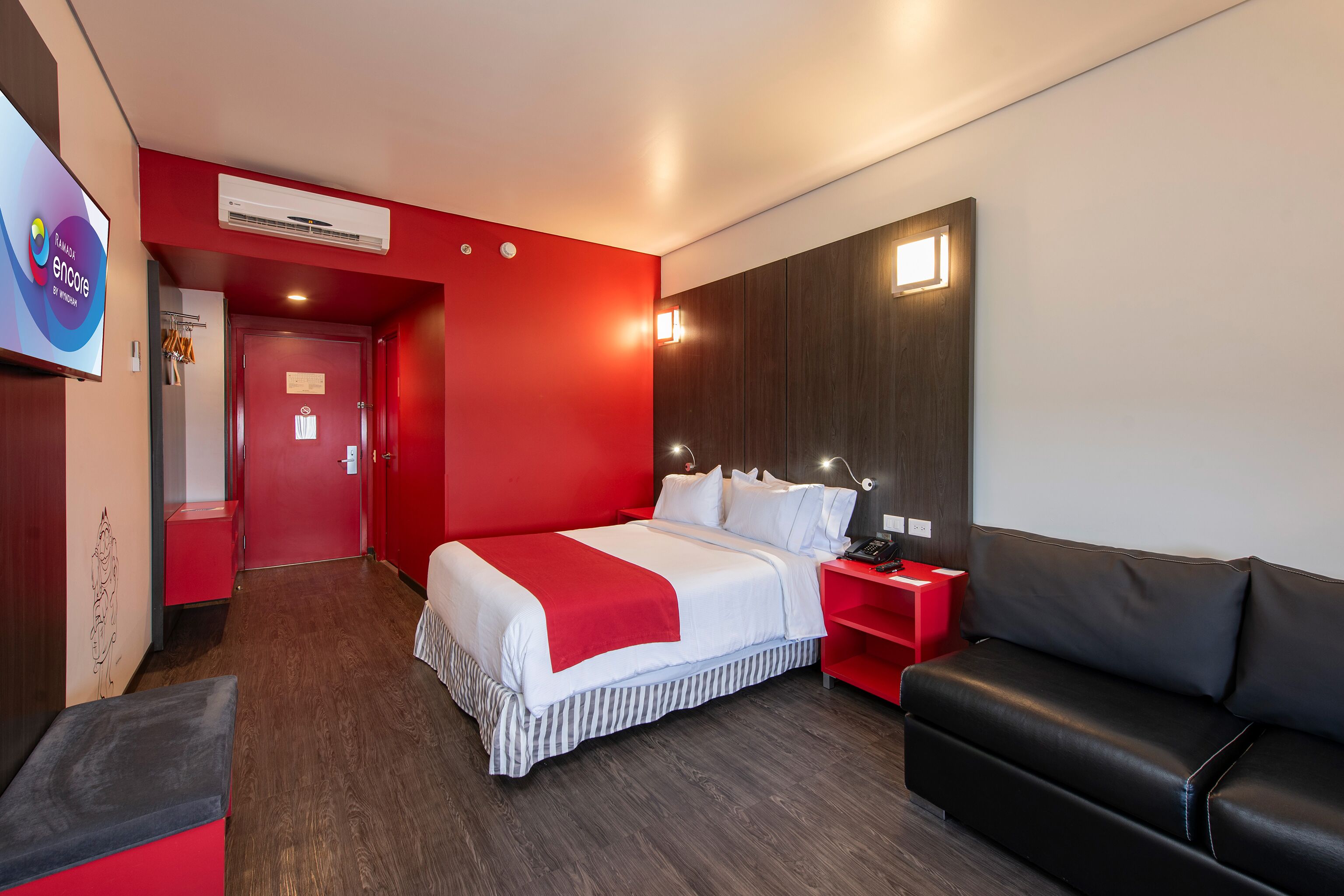 A hotel room with a large bed, a red wall, a black leather sofa, and a red nightstand.