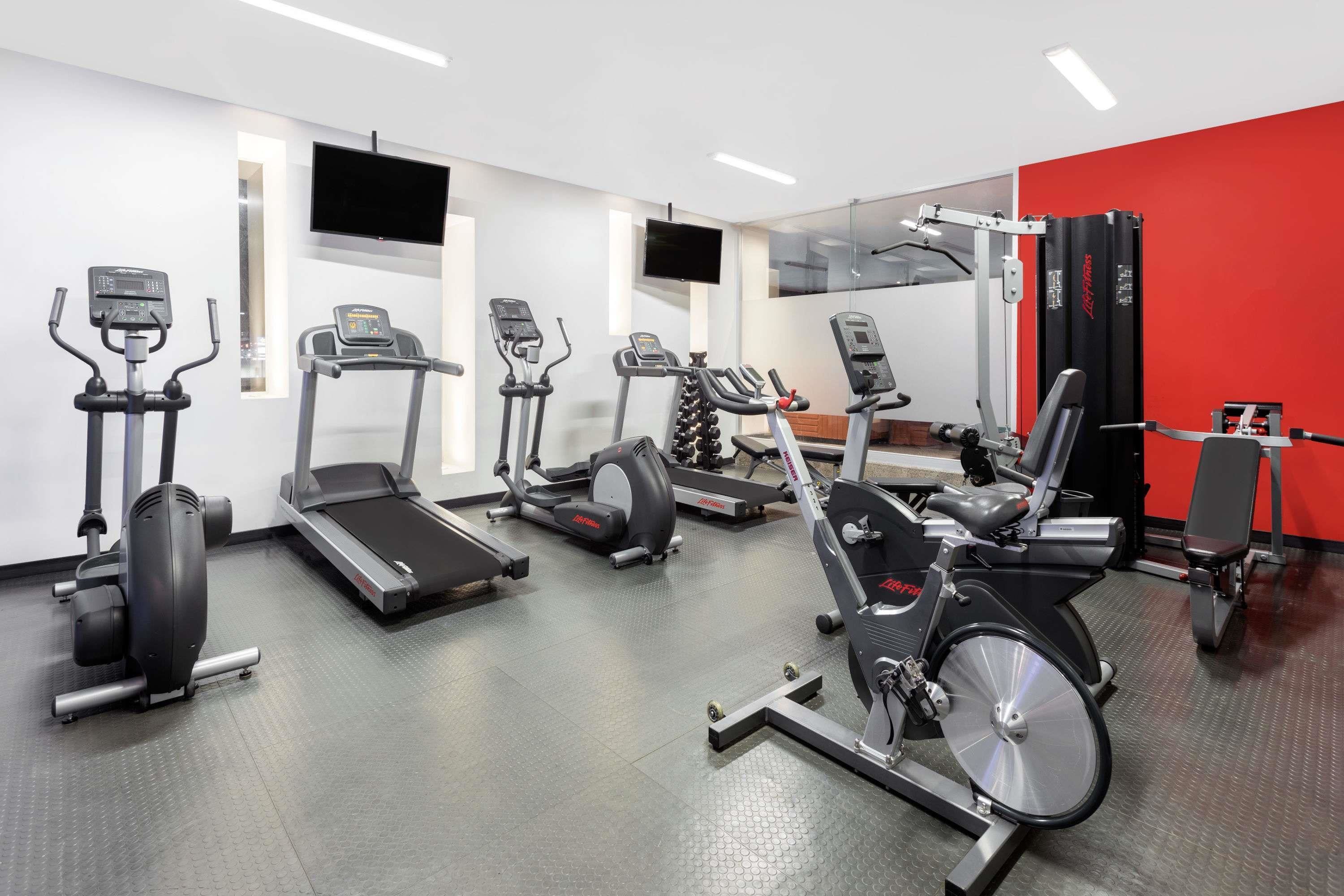 Un gimnasio con diversas máquinas de cardio, un televisor y una pared de acento roja.