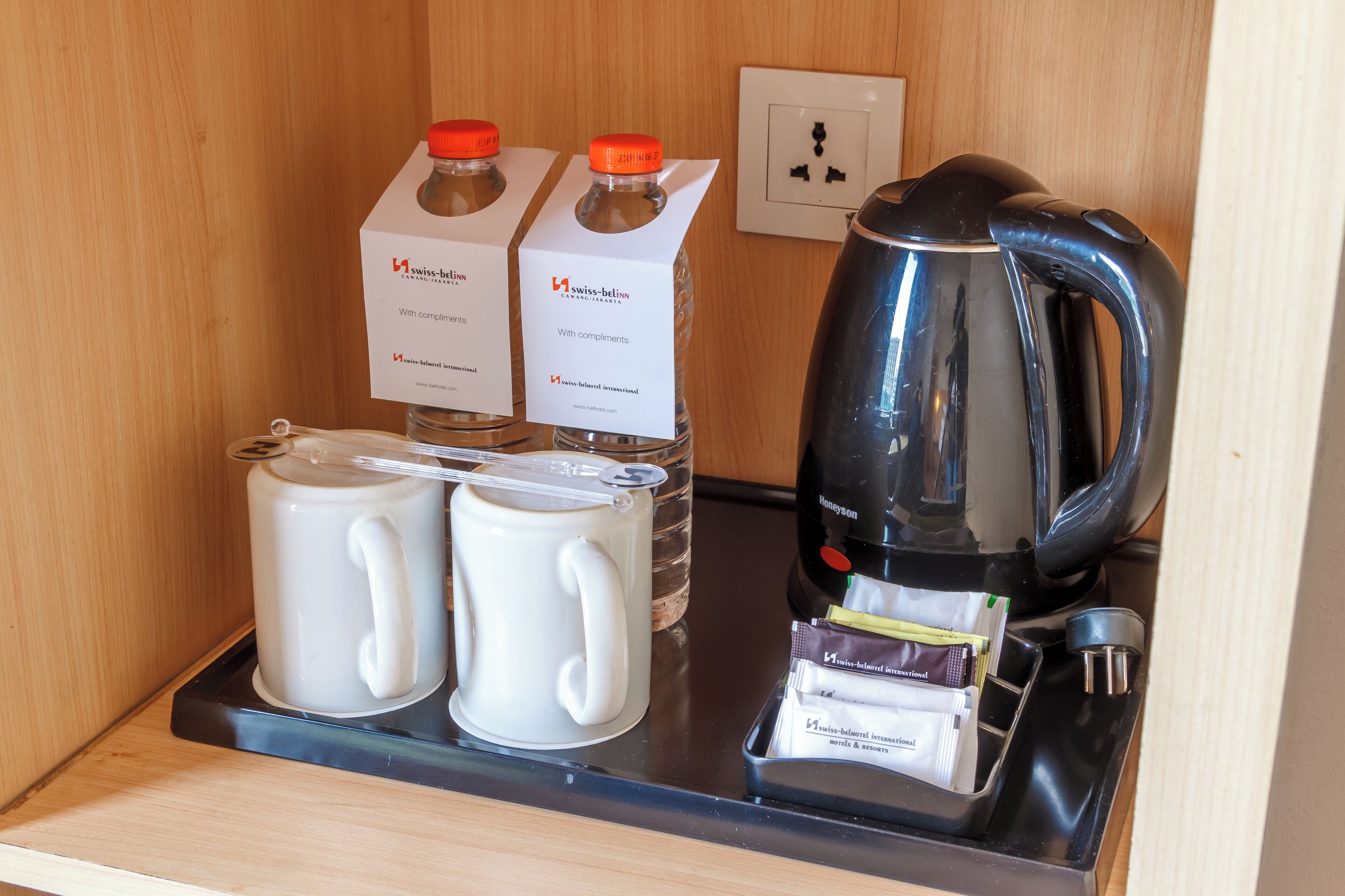A hotel minibar with two milk cartons, two white mugs, a black coffee maker, and a tray with various items.