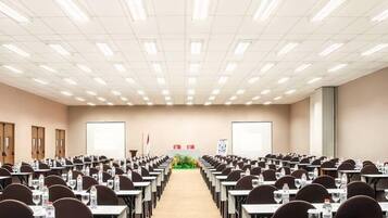 A conference room with rows of tables and chairs, equipped with water bottles and glasses, and a projector screen at the front.