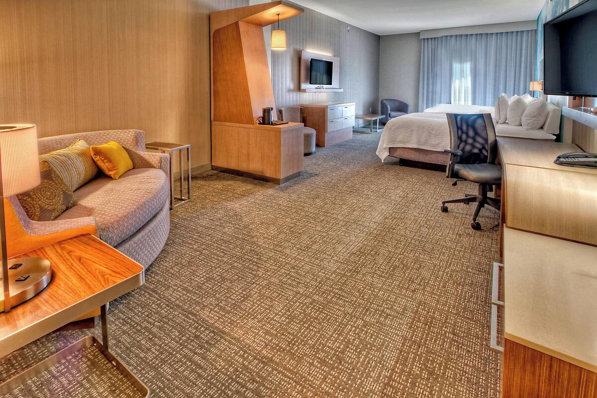 A hotel room with a sofa, a desk, a bed, a TV, and a desk lamp.
