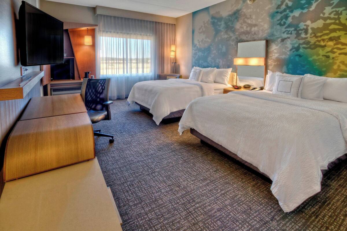 A hotel room with two beds, a desk, a TV, and a large window with curtains.